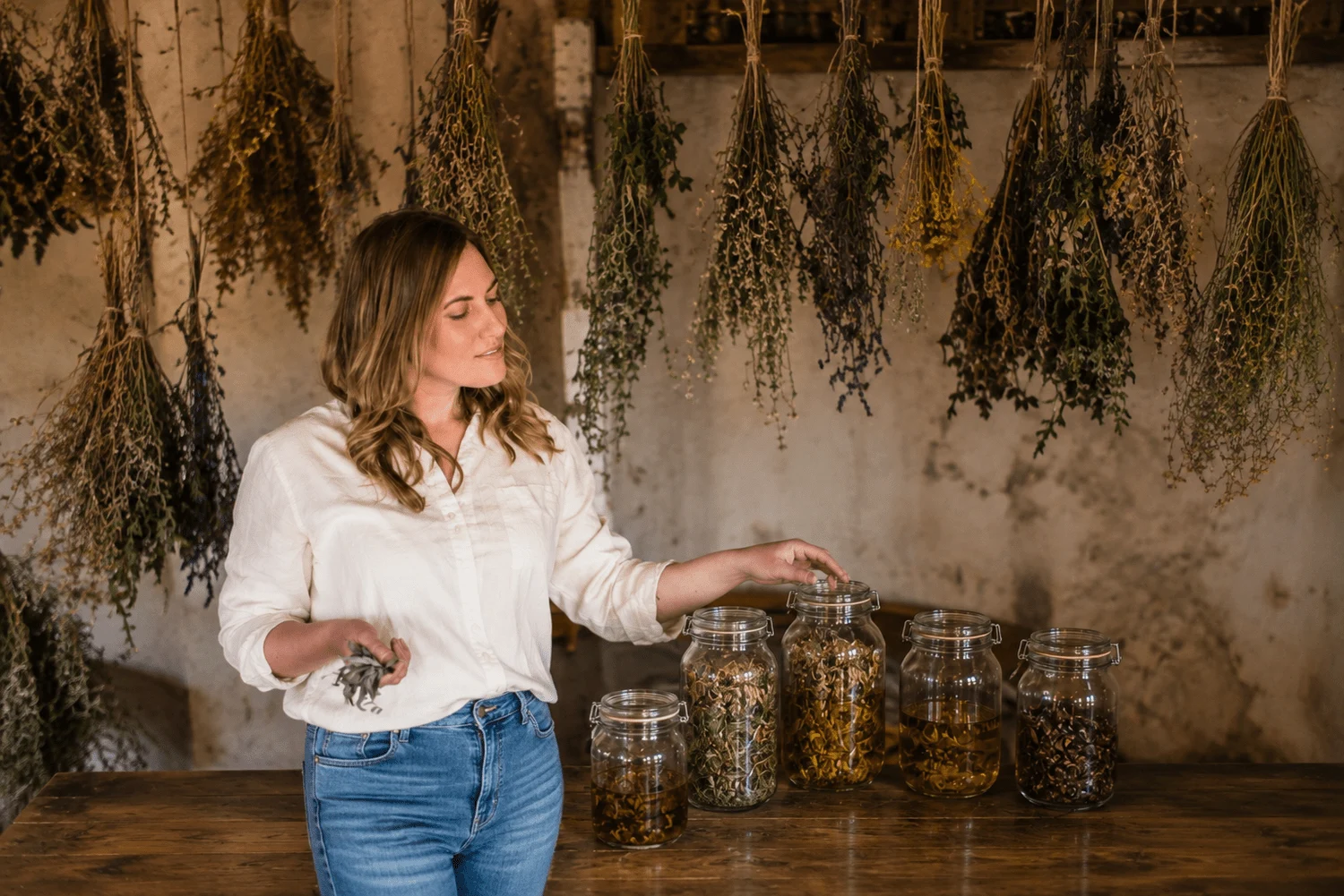 A woman with wavy, shoulder-length hair wearing a white shirt and blue jeans stands at a wooden table. She is holding a small branch in her left hand and opening a large jar with her right hand. On the table are five glass jars filled with various dried herbs or flowers. Behind her, dried herbs hang from the ceiling against a rustic wall.