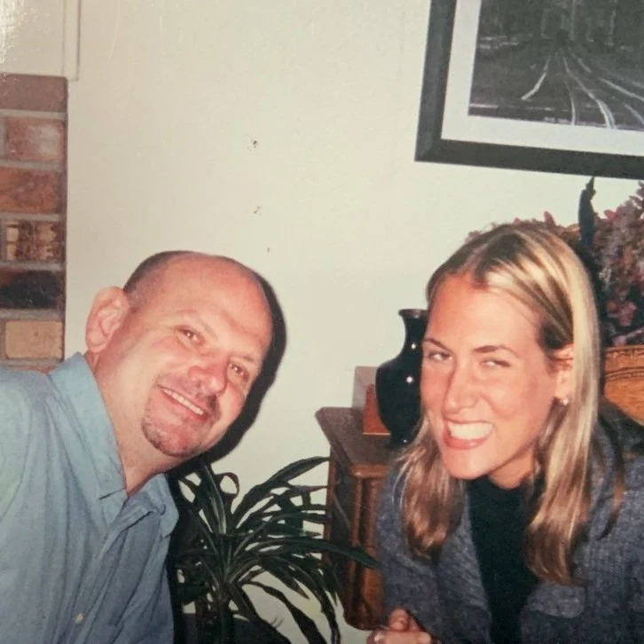 A man and woman sitting indoors, smiling for a photo, with houseplants and framed art in the background.