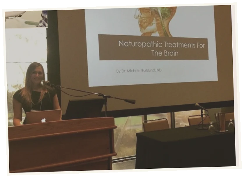 A woman standing at a podium with a laptop, giving a presentation on 'Naturopathic Treatments For The Brain' by Dr. Michele Burklund, ND, with a projected slide showing a brain illustration.