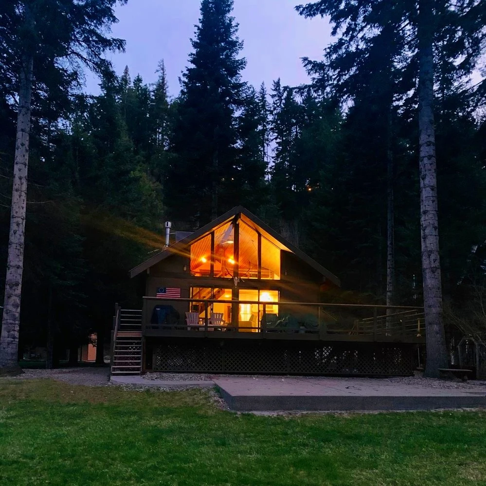 A cozy cabin with large glass windows and a front porch illuminated with warm lights, surrounded by tall trees in a forest setting during evening