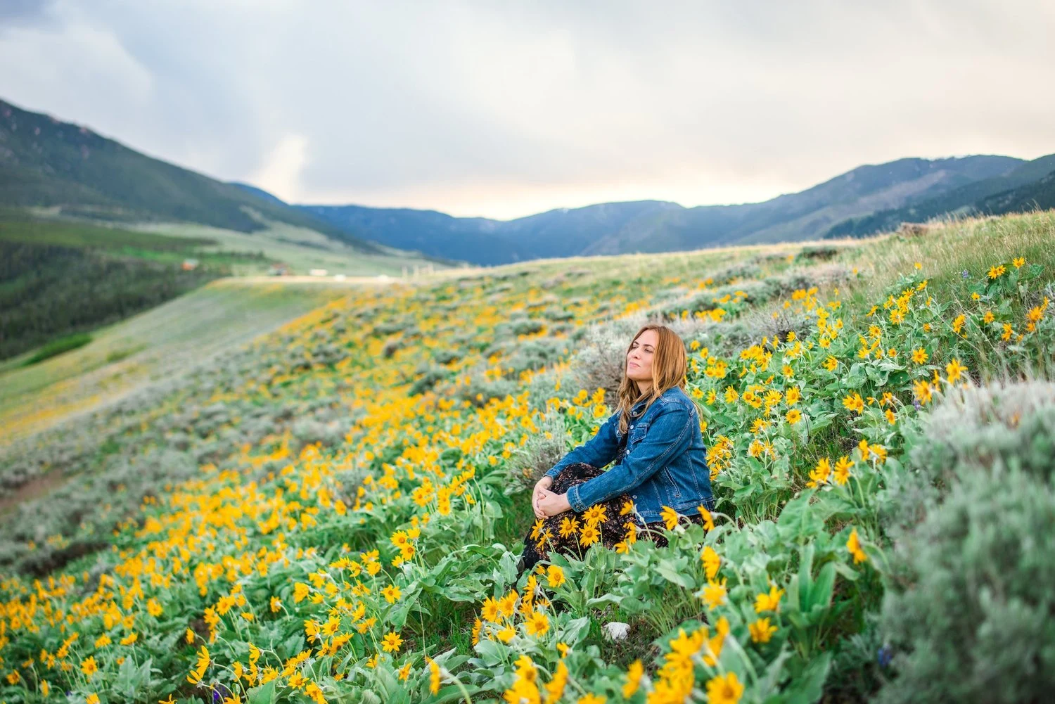 A woman with long hair sitting among yellow flowers in a green field with mountains and cloudy sky in the background.